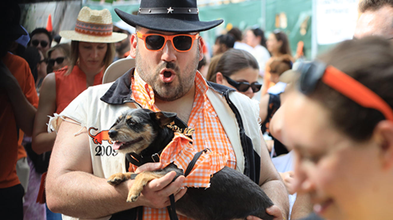 A man in the P-rade wears orange sunglasses and a black cowboy hat and carries a dog.