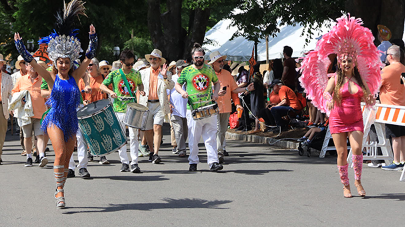 People marching in the P-rade bang drums and wear feathered headdresses.