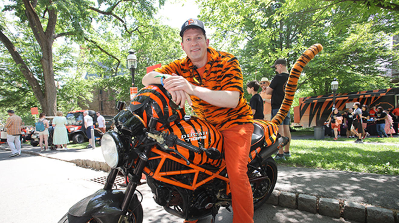 A man in a tiger striped shirt and orange pants leans on a motorcycle covered with tiger stripes that has a long fuzzy tiger tail.