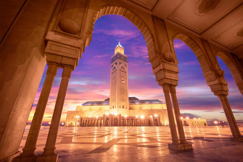 Tiger-Travels_Casablanca-Morocco-Alaoui.jpeg A view between pillars looks out to the Hassan II Mosque. The tall structure is surrounded by the pink and purple skies of sunset.