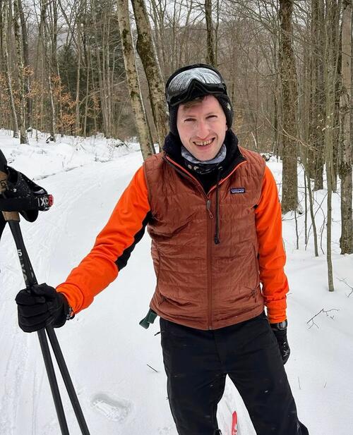 The author, Harrison Blackman ’17, stands outside in the snow in an orange shirt and puffer vest with ski gear