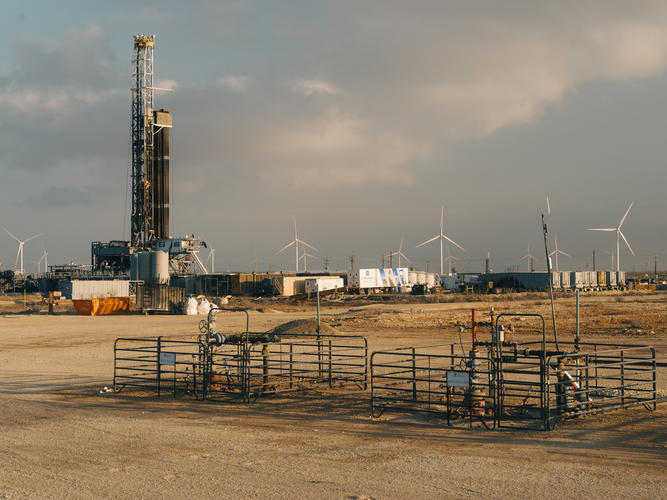 A large rig is visible in the background. Further back are several white wind turbines. In the foreground we see two metal fences around pipes.