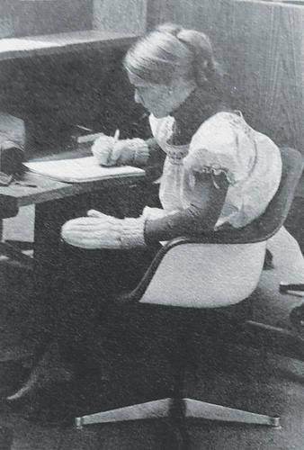 Black and white photo of a woman writing at a desk while wearing woolen mittens.
