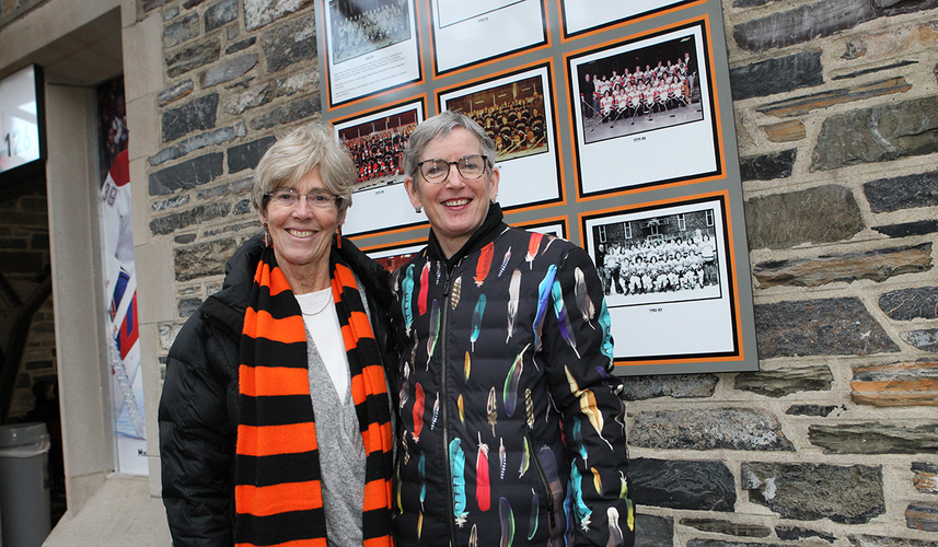 Carol Brown ’75, left, and Elizabeth English ’75 stand in front of a wall of hockey team photos on a stone wall at Baker Rink.