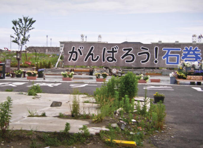 A brightly painted sign in a heavily damaged area reads, “Let’s do our best – we can do it, Ishinomaki!”