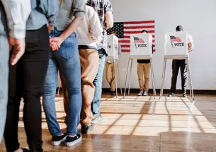 A line of people waiting to vote is shown from behind, from the waist down. People vote in the background.