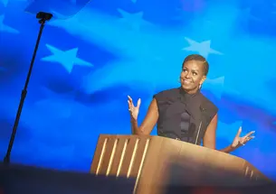 Michelle Obama ’85 speaks at a podium in front of a blue starred background; a teleprompter is in the foreground.
