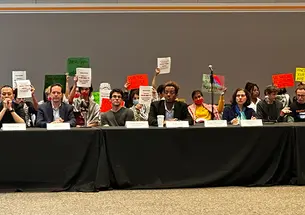 Pro-Palestinian students sit behind members of the CPCU's table holding signs.