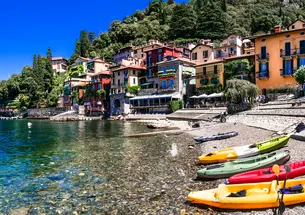 The edge of Lake Como in Italy, with clear water showing pebbles at the bottom, colorful plastic canoes lined up in the foreground, and charming brightly colored Italian homes in the background.