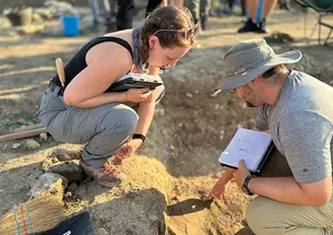 Two people at an archaeological dig examine something in the dirt.