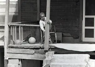 In this black-and-white photo from the 1960s, a boy stands on the porch of a home in rough shape, talking to another boy on the ground.