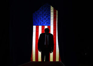 Trump standing in front of a lit U.S. flag surrounded by darkeness