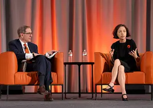 Two people sit on orange chairs on a stage.
