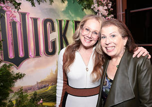 Two women smile for the camera in front of a poster for "Wicked" the movie.