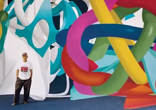  Frank Stella wearing a Make Art Not War t-shirt standing in front ofa huge colorful painting