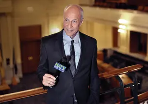Michael Aron *70 holds a microphone while speaking from an upper level of the NJ statehouse.