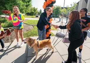 President Eisgruber shakes a woman's hand in front of an orange-and-black balloon arch; his other hand holds the leash of a yellow labrador.