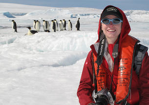 A woman in a red jacket and sunglasses holds a camera in an ice-covered place, while about a dozen emperor penguins stand in the background.