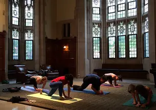 Five people practice yoga in a dim room with Gothic windows.