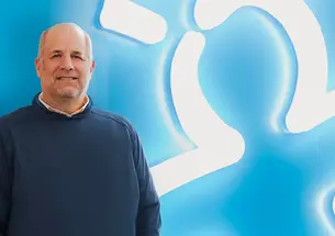 Rick Woldenberg ’81 against a blue background where his company's logo — a person with lines like a lighbulb overhead — is glowing in white neon bulbs.