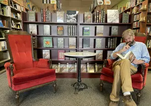 John Kuhner sits at one of two red chairs and reads a book in front of a bookcase in his bookstore.
