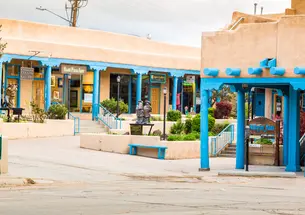 A view McCarthy Plaza in Taos, New Mexico which has Pueblo style buildings with bright blue trim around the stores.