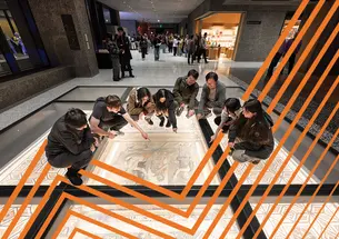 Students gather around a floor mosaic under glass. Orange zigzags reminiscent of the Venture Forward campaign logo are superimposed over the image.