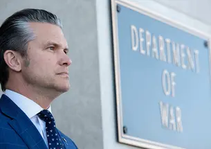 Pete Hegseth stands outside in front of a sign on a building reading "Department of War."