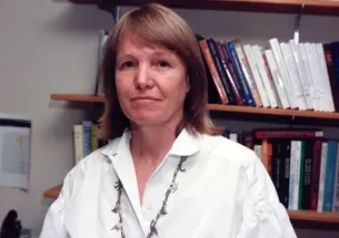 Portrait of Kay Warren standing in an office or study, wearing a white blouse and silver necklace, with bookshelves filled with books in the background. She faces the camera with a calm, neutral expression.