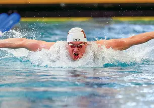Mitchell Schott competing in a butterfly race in an indoor swimming pool, wearing a swim cap and goggles as water splashes around him during a powerful stroke.