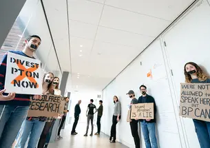 A few students line a hallway holding signs that say "Divest PTon now" and "We're not allowed to speak here but BP can."