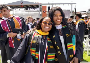 Two women smile for the camera at a graduation ceremony, wearing multicolored stoles and cords over their black graduation robes.