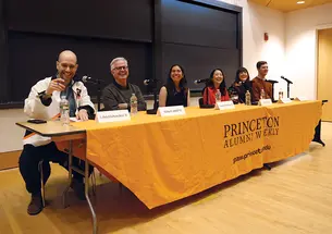 Six people sit at a table in the front of a classroom; the table has an orange tablecloth that reads Princeton Alumni Weekly.