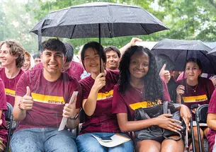 Students flash thumbs up from under black umbrellas in the rain.