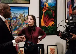Wearing an audio kit, Lydia Cornett looks at a man who's speaking to her; next to them another person holds a technical setup that could be a camera.