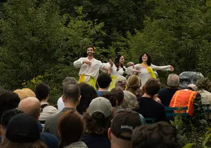 Three actors perform on a stage outside before a small audience.
