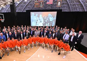 A large group of alumni stand behind the current men's basketball team, who are on one knee. In the background is a large screen showing photos of Pete Carril.