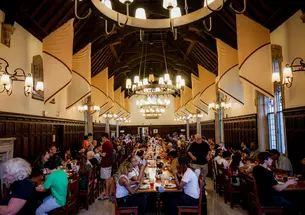 Students sit at long tables arranged in three rows in a dining hall with a high wood ceiling.