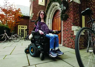 This is a photo of Naomi Hess ’22 in her motorized wheelchair in front of a Princeton campus building. Bicycles are in the foreground and background.