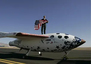 Brian Binnie *78 stands on what looks like a small airplane, holding an American flag.