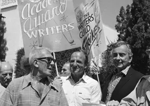 Black-and-white photo of three men in a crowd holding signs reading "Academy Award writers" and "The Writers Guild Strikes."