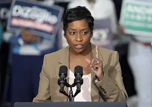Andrea Campbell speaks at a podium; people hold campaign signs in the background.