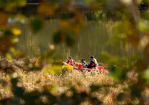 Seen through an opening in the trees, two people in orange vests motor on Lake Carnegie in an orange boat.