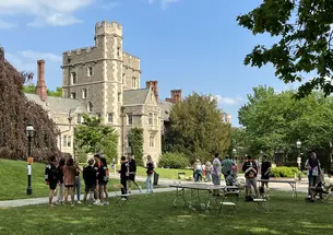Students socialize by tables in a courtyard, with a stone Gothic building in the background..