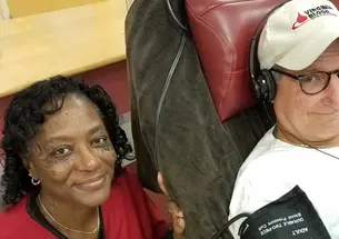 Wearing a "Virginia Blood" baseball cap, Doug Schutte ’76 sits in a chair wearing headphones and a blood pressure monitor as a Red Cross worker poses next to him.
