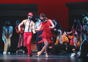 This is a photo of students on stage during a Triangle Club rehearsal. A boy and a girl dance in front while others snap their fingers while watching in back and on the sides. All are wearing black KN95 masks.