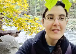 Elizabeth Tsurkov takes a selfie under a tree with a small river behind her.
