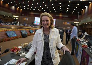 Isobel Coleman ’87 faces the camera and smiles in a room where each seat has a small screen. Blurry in the background are lit-up signs reading G20.