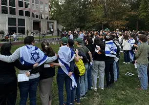 People stand with their arms around each other's shoulders outside a campus building, listening to two students play music. A few have blue-and-white Israel flags wrapped around their shoulders.