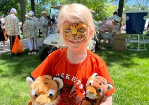 A six-year-old girl with tiger faceprint, an orange shirt, and a plush tiger under each arm.